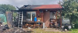 Dom letniskowy drewniany, po lewej stronie spalony, o dom oparta aluminiowa drabina, po prawej stronie drzewo 
