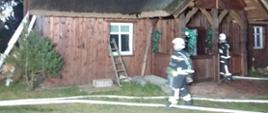 Drewniany dom ze strzechą. Wokół domu strażacy uczestniczący w akcji gaszenia. Widać 2 linie gaśnicze.
