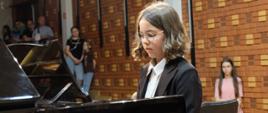 Dziewczynka w okularach gra na fortepianie, w tle oparta o ceglaną ścianę publiczność.