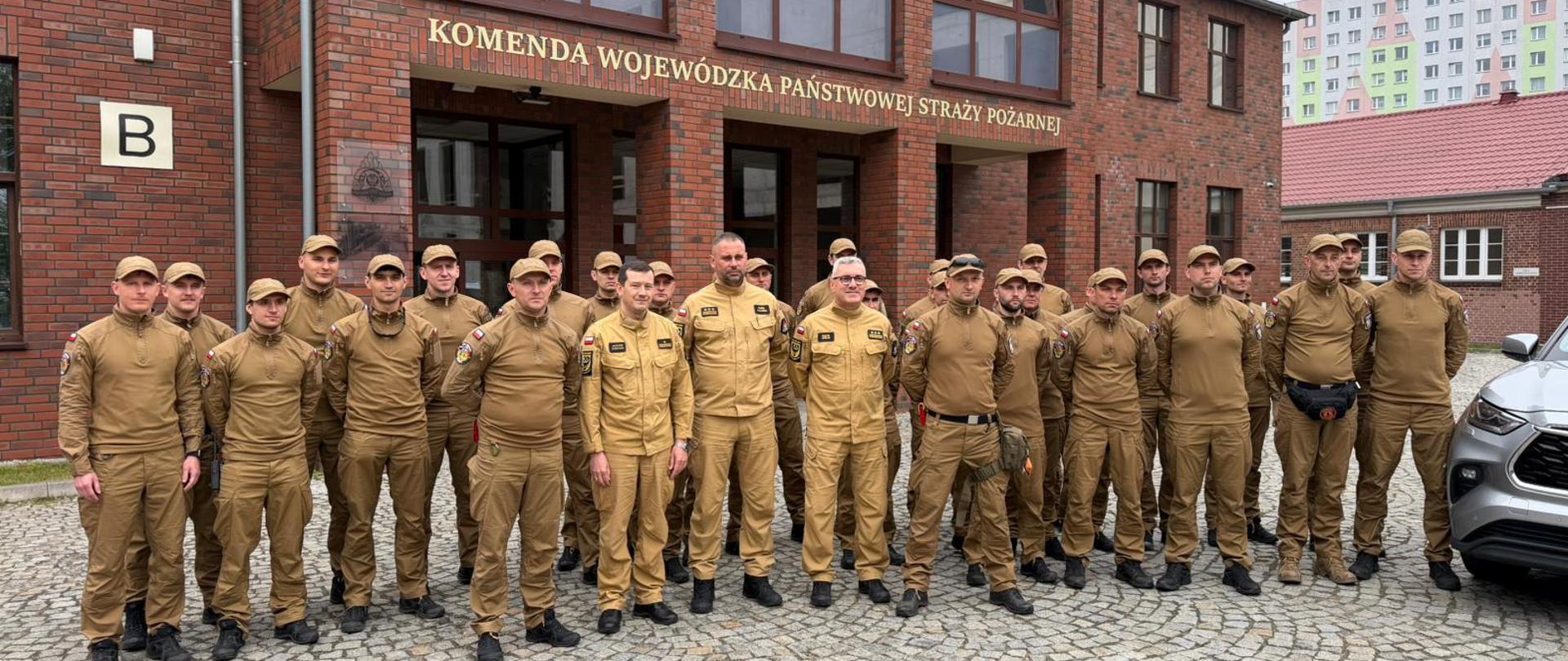  Zapytanie zostało przetworzone
Zdjęcie przedstawia grupę ponad dwudziestu strażaków w piaskowych mundurach służbowych, pozujących do wspólnej fotografii przed budynkiem komendy
