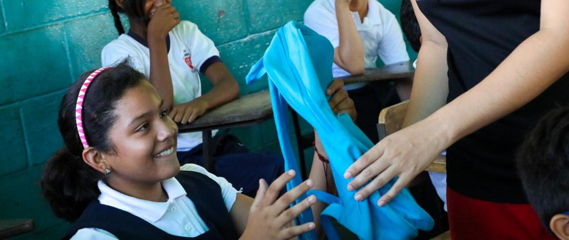 W klasie uśmiechnięta dziewczynka odbiera z rąk nauczycielki prezent