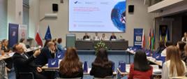 Sesja plenarna uczestników i organizatorów programu - kilkanaście osób siedzi przy stołach ustawionych w podkowę; na dalszym planie przedstawiciele organizatorów siedzą przy stole prezydialnym, nad nimi ekran multimedialny
