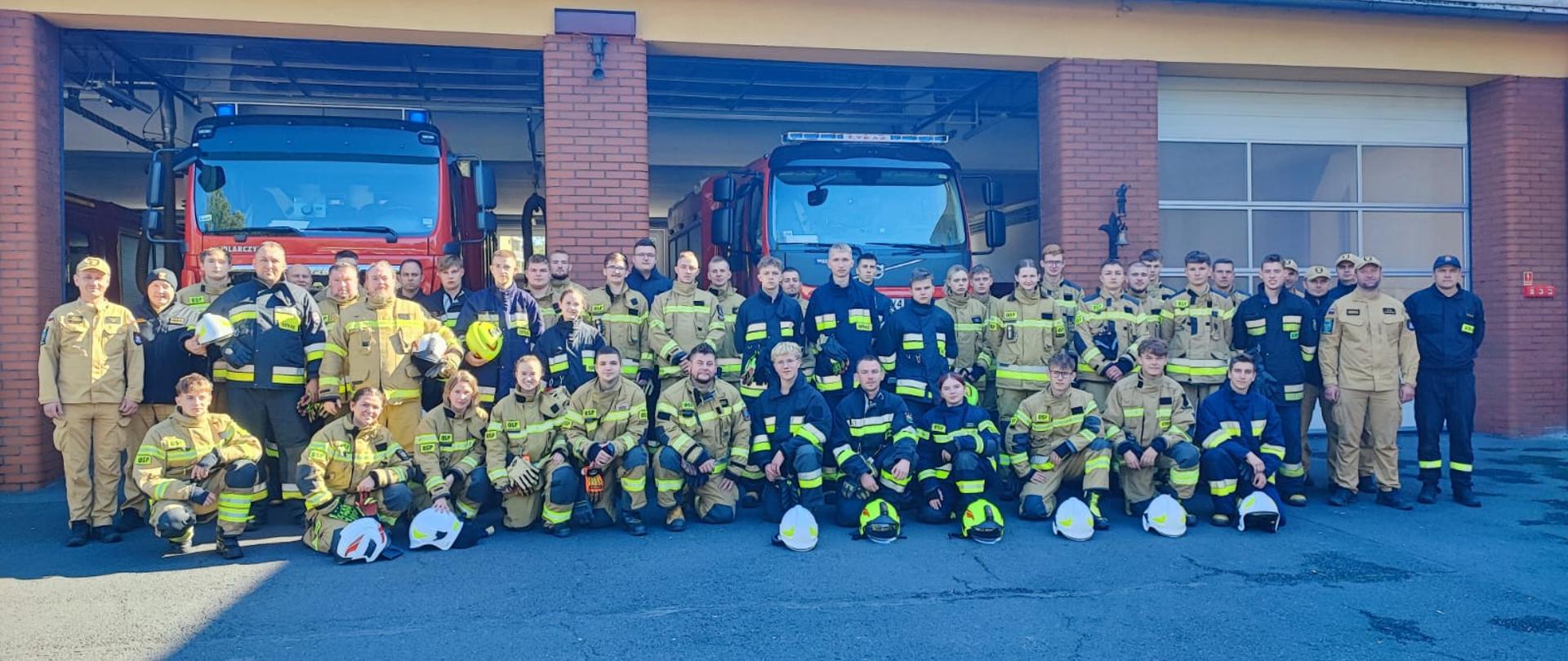 Na zdjęciu znajdują się absolwenci szkolenia podstawowego strażaka ratownika OSP oraz kadra szkoleniowa. Kursanci ubrani w ubrania specjalne. Zdjęcie zrobione na tle bram garażowych JRG w Oleśnie i samochodów pożarniczych.