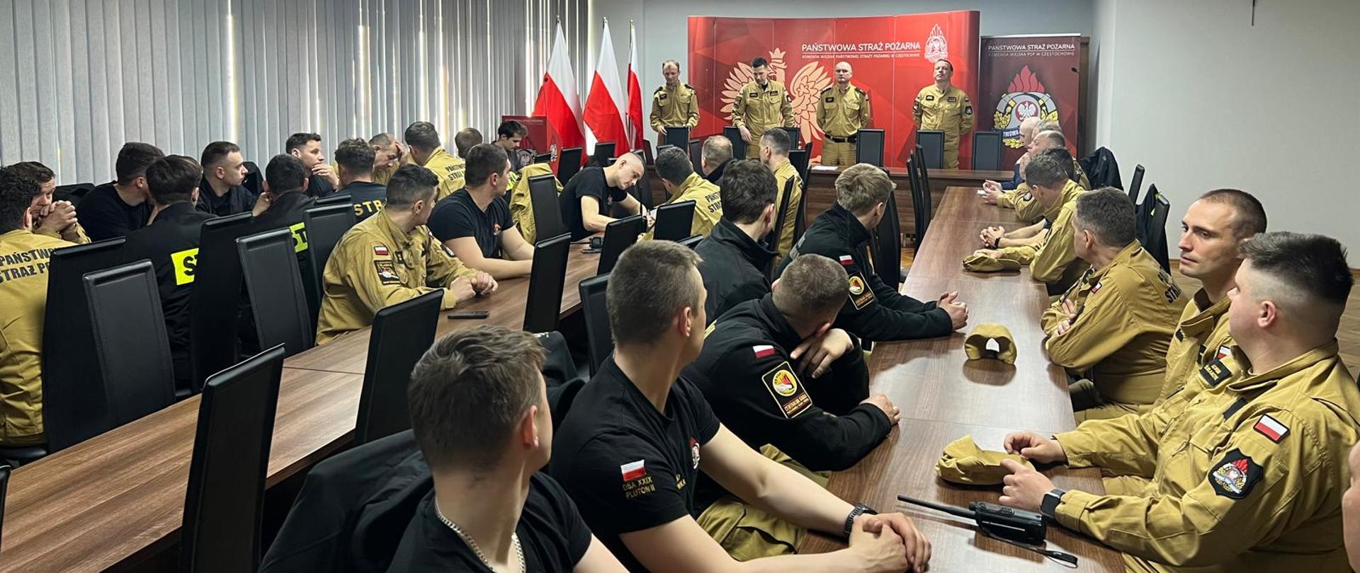 To zdjęcie przedstawia formalne spotkanie lub odprawę strażaków w sali konferencyjnej. Przy długich stołach siedzi kilkudziesięciu uczestników – część w mundurach służbowych w kolorze piaskowym, inni w czarnych koszulkach z oznaczeniami. Wszyscy są zwróceni w stronę przodu sali. Na czele pomieszczenia stoi kilku przełożonych w mundurach, przemawiających do grupy. Za nimi widać czerwone tło z emblematem straży pożarnej oraz napis „Państwowa Straż Pożarna”. Obok ustawione są flagi Polski.