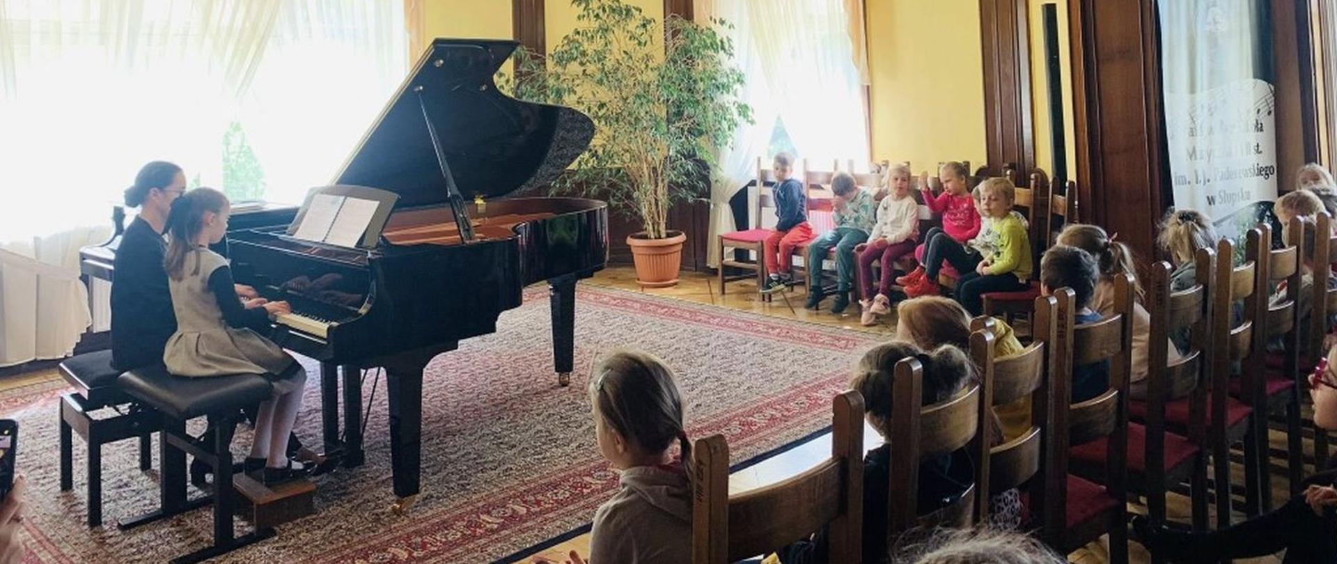 Zdjęcie kolorowe. W eleganckim wnętrzu sali kameralnej przy fortepianie siedzi mała dziewczynka ze swoją nauczycielką, grają w duecie z nut, które znajdują się na pulpicie fortepianu. Przed nimi oraz po prawej stronie na stylowych krzesłach siedzi zasłuchana publiczność. Wysoko, na żółto - brązowych ścianach umieszczone jest godło Polski. Atmosfera zdjęcia jest pełna skupienia.