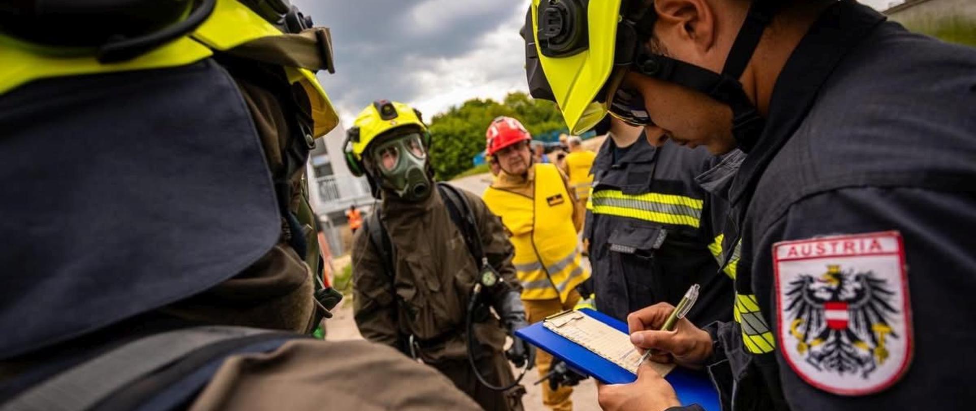 Na zdjęciu strażacy różnych narodowości, w różnych mundurach, w żółtych kaskach, na pierwszym planie plakietka z napisem AUSTRIA