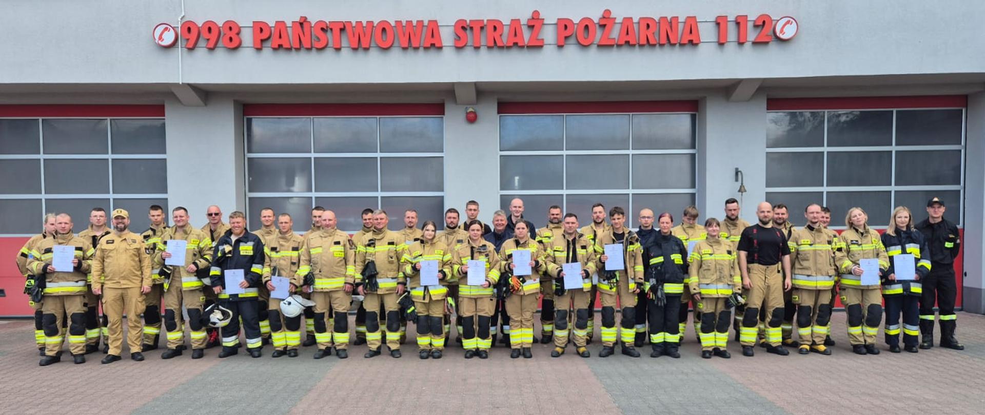Uczestnicy szkolenia podstawowego strażaka ratownika OSP po zakończeniu kursu, ustawieni do wspólnego zdjęcia przed budynkiem Komendy Powiatowej PSP w Przasnyszu.