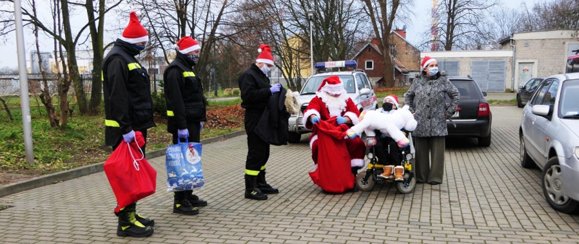 Czworo strażaków na parkingu samochodowym przed blokami wręcza prezenty Angelice, troje z nich jest w ubraniach koszarowych, jeden jest przebrany za Mikołaja. Dziewczynka siedzi na wózku. Na zdjęciu znajduje się także jej mama.