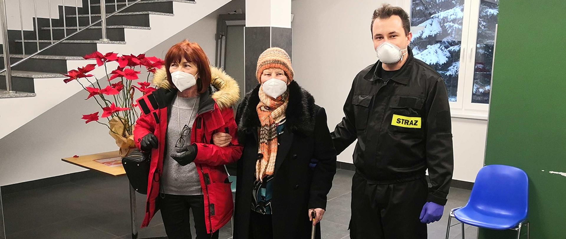 Na zdjęciu widać seniorkę wraz z swoją opiekunką i strażakiem w punkcie szczepień.