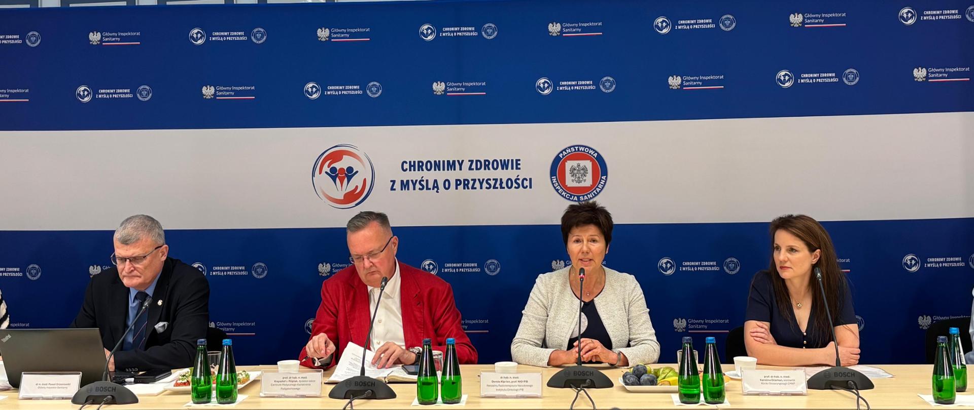 Panel dyskusyjny z czterema osobami siedzącymi przy długim stole na tle niebiesko-białej ścianki z logotypami instytucji państwowych. W tle widoczne jest powtarzające się logo Głównego Inspektoratu Sanitarnego oraz hasło
„CHRONIMY ZDROWIE Z MYŚLĄ O PRZYSZŁOŚCI” umieszczone centralnie na białym pasie, obok okrągłych emblematów graficznych. Przy stole siedzą cztery osoby, od lewej do prawej: mężczyzna o krótkich siwych włosach w ciemnym garniturze, patrzący w dół na laptop; mężczyzna w białej koszuli i czerwonej marynarce, zajęty dokumentami; kobieta z krótkimi, ciemnymi włosami, w jasnej marynarce, mówi do mikrofonu stojącego przed nią; młodsza kobieta o długich, brązowych włosach w granatowej sukience, z założonymi rękami, przysłuchuje się.
Na stole przed każdym uczestnikiem leżą mikrofony konferencyjne marki Bosch, kartki z dokumentami i tabliczki z imieniem, nazwiskiem i funkcją, których tekst jest zbyt mały, aby dało się go odczytać. Na stole stoją również zielone szklane butelki z wodą, plastikowe kubki oraz talerzyki z drobnymi przekąskami i owocami, w tym talerzyk ze śliwkami i cytrynami.