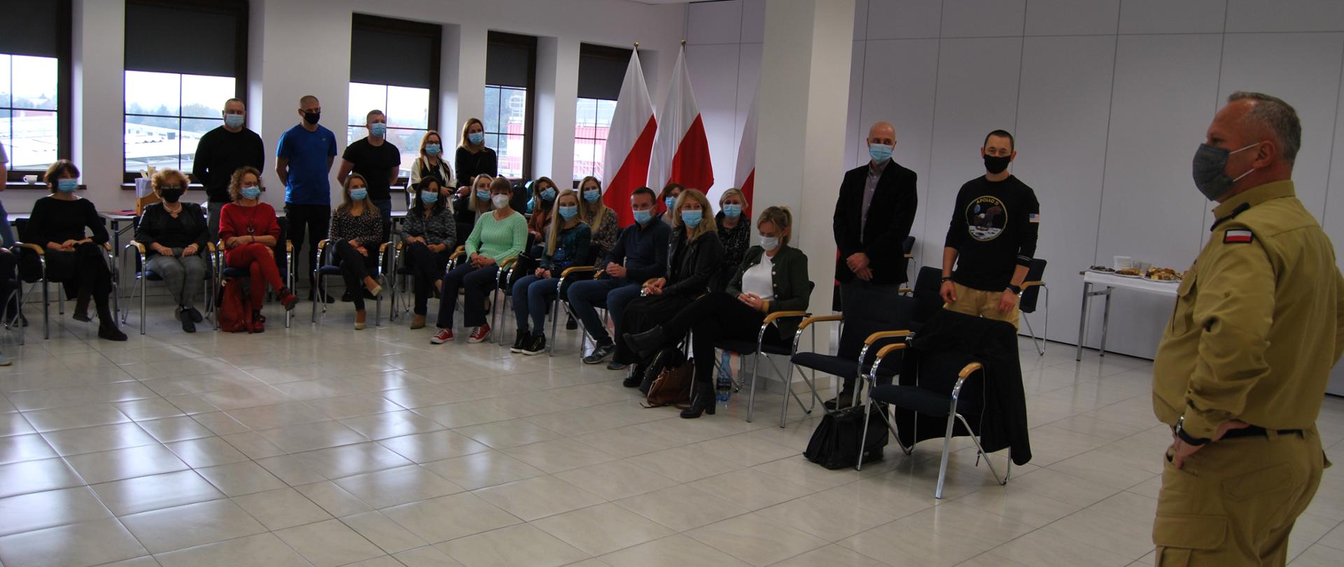 Na zdjęciu widać siedzących oraz stojących w półokręgu mężczyzn oraz kobiety przemawia do nich stojący na środku zastępca kujawsko-pomorskiego komendanta wojewódzkiego PSP, na twarzach mają założone maseczki. Zdjęcie wykonano w sali konferencyjnej Ośrodka Szkolenia KW PSP w Toruniu z siedziba w Łubiance