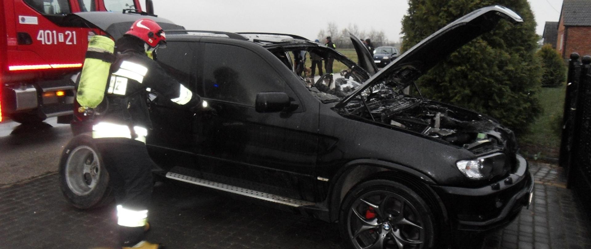 Zdjęcie przedstawia strażaka w ubraniu specjalnym działającego przy samochodzie osobowym, w tle samochód pożarniczy.