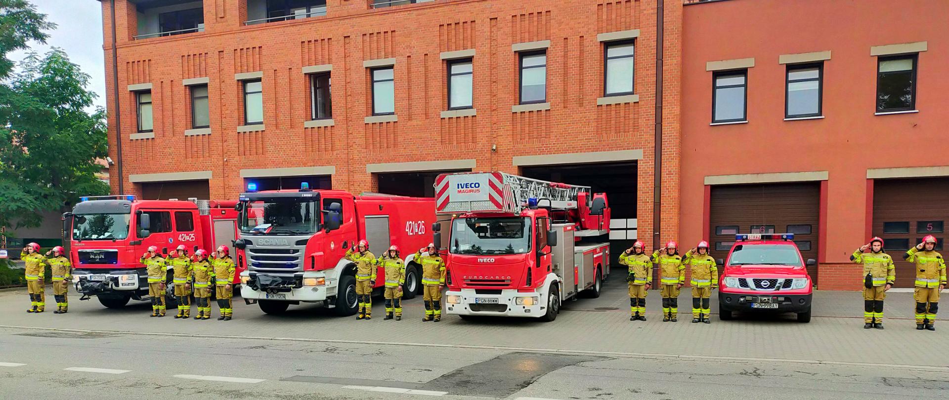 Zdjęcie zrobione przed strażnicą. Na zdjęciu widać cztery samochody strażackie, między pojazdami stoją strażacy w ubraniach bojowych i salutują oddając cześć bohaterom biorącym udział w walce o Niepodległą Polskę