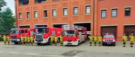 Zdjęcie zrobione przed strażnicą. Na zdjęciu widać cztery samochody strażackie, między pojazdami stoją strażacy w ubraniach bojowych i salutują oddając cześć bohaterom biorącym udział w walce o Niepodległą Polskę