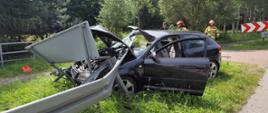 Rozbity samochód osobowy, który uderzył w barierę energochłonną. Przebiła ona przednią szybę pojazdu. W tle widać strażaków.