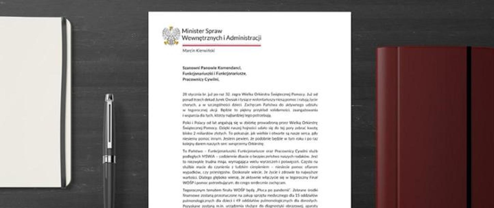 Zdjęcie przedstawia leżący na biurku list ministra spraw wewnętrznych i administracji skierowany do podległych ministerstwu służb dotyczący współpracy z wielką orkiestrą świątecznej pomocy. Po lewej stronie na biurku znajduje się czarne pióro natomiast po prawej brązowy notes. 