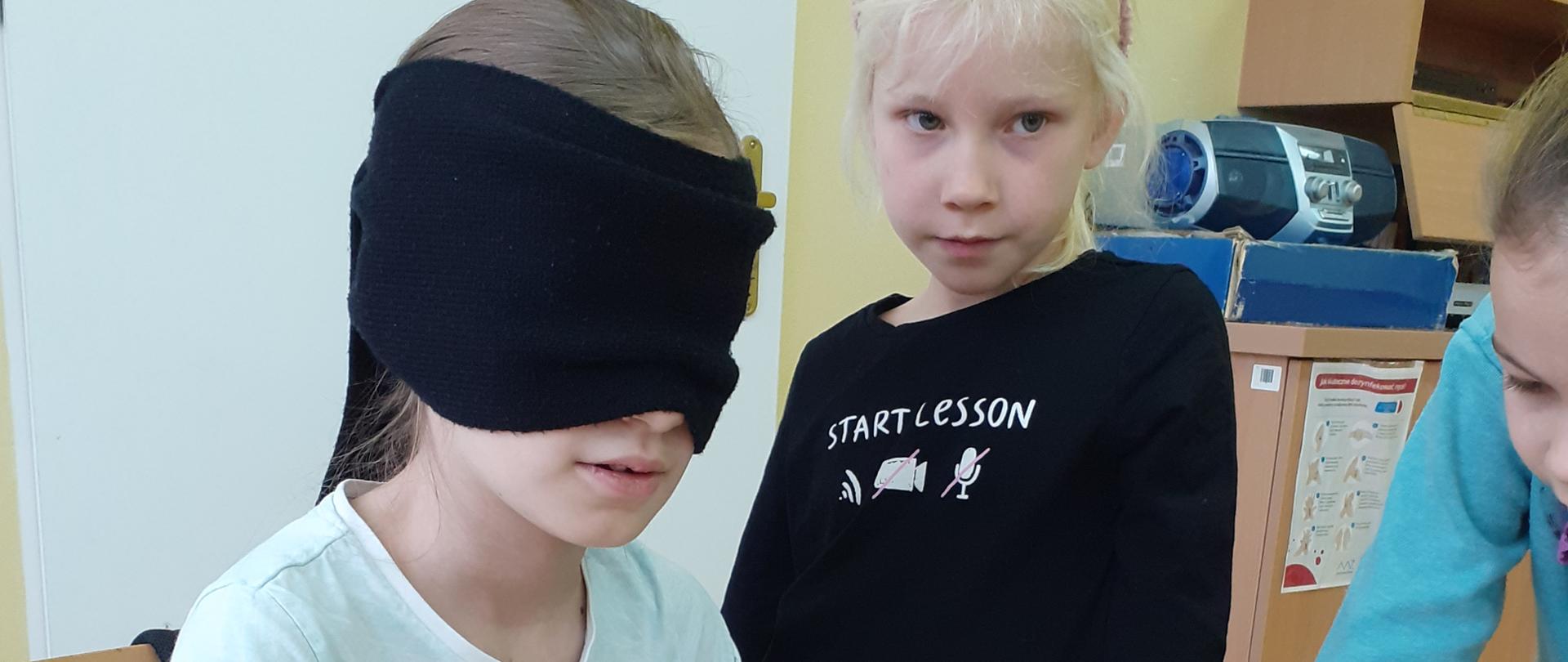Zdjęcie dziecka w świetlicy szkolnej z zasłoniętymi oczami, siedzącego przy ławce, próbujące coś napisać. Obok stoją dzieci.