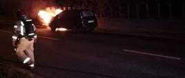 Na zdjęciu widzimy palący się samochód oraz strażaka podczas przygotowania do działań. Zdjęcie wykonane w porze nocnej. Samochód pali się na jezdni na prawym pasie drogi.