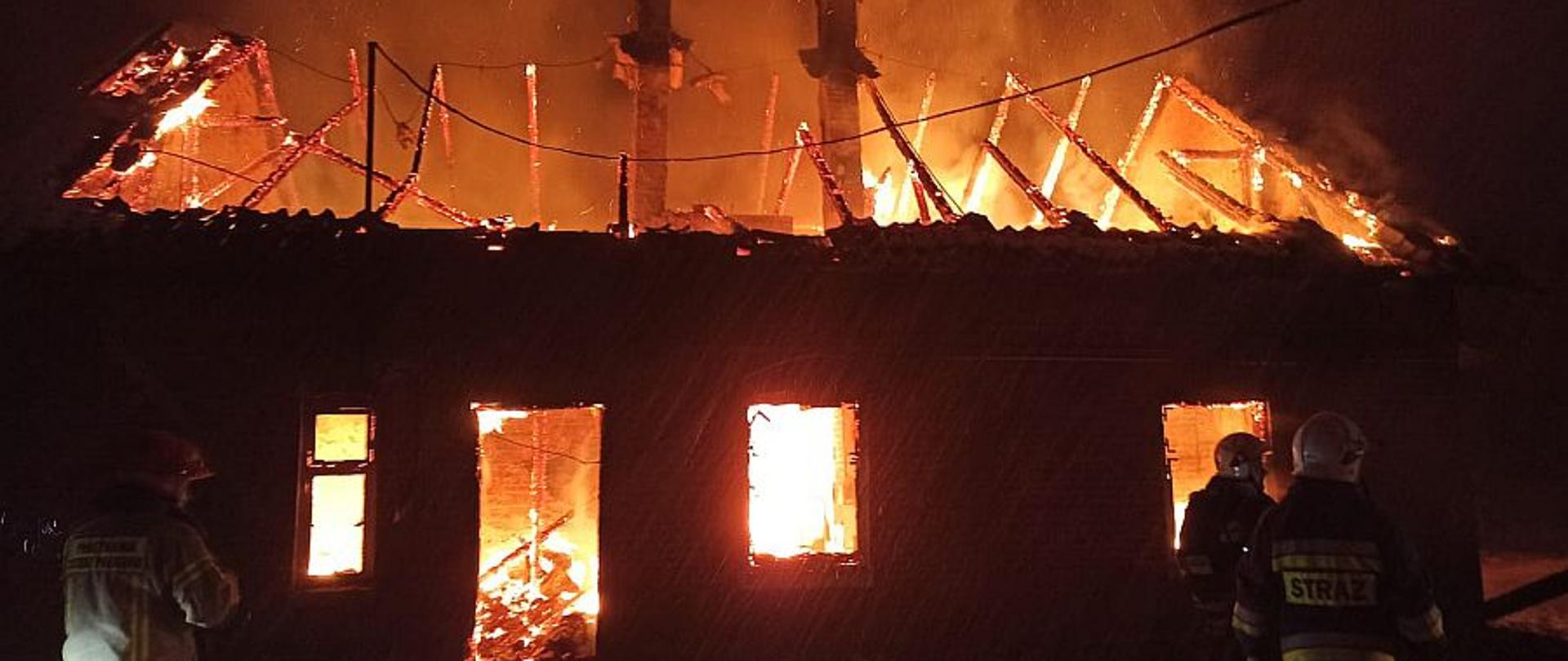 Płonący budynek mieszkalny z zawalonym dachem, w środku wystają dwa kominy. Dookoła stoją strażacy w ubraniach specjalnych i hełmach. Płomienie są widoczne u góry oraz w otworach okiennych i drzwiach na parterze. 