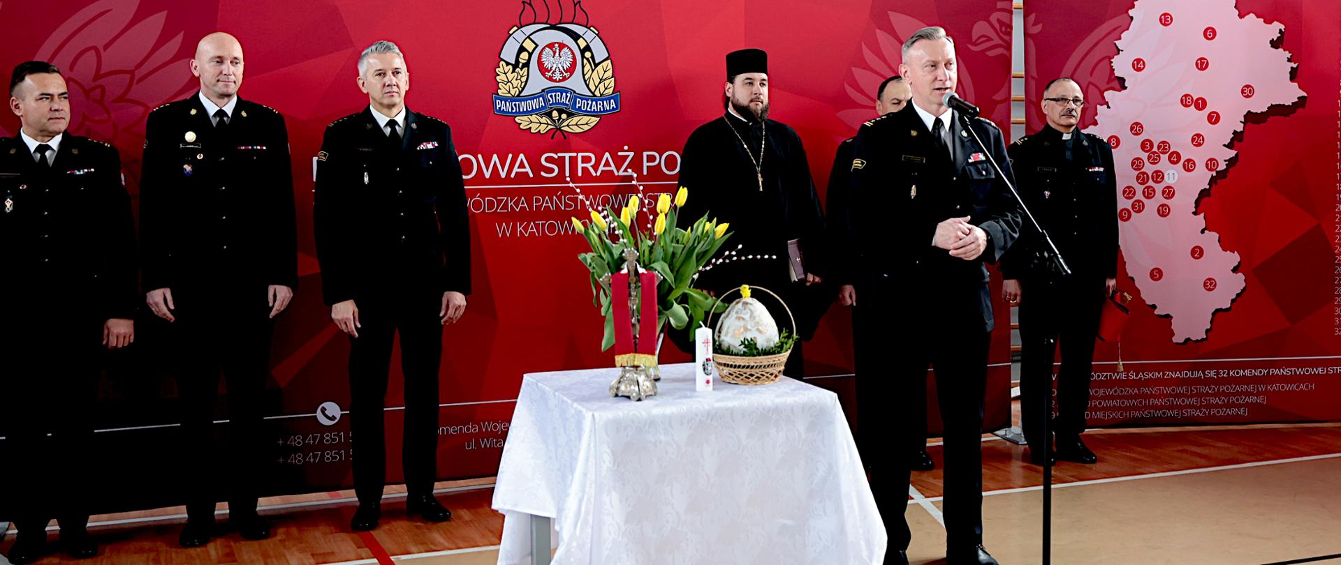 Na zdjęciu widać uroczyste spotkanie świąteczne w sali sportowej Komendy Wojewódzkiej Państwowej Straży Pożarnej w Katowicach. Na tle czerwonej ścianki z dużym emblematem straży stoi kilku mężczyzn w czarnych, wyjściowych mundurach strażackich są to zastępcy śląskiego komendanta wojewódzkiego. Po prawej stronie śląski komendant wojewódzki starszy brygadier Damian Legierski przemawia do mikrofonu. Obok niego stoją inni uczestnicy, w tym duchowni w czarnych strojach. Na środku, przed nimi, ustawiony jest stół przykryty białym obrusem. Leżą na nim świąteczne symbole: świeca, kwiaty oraz koszyk z pokarmami. W tle, po prawej stronie na ściance, widoczna jest mapa z zaznaczonymi punktami i lista jednostek straży. 