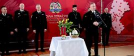Na zdjęciu widać uroczyste spotkanie świąteczne w sali sportowej Komendy Wojewódzkiej Państwowej Straży Pożarnej w Katowicach. Na tle czerwonej ścianki z dużym emblematem straży stoi kilku mężczyzn w czarnych, wyjściowych mundurach strażackich są to zastępcy śląskiego komendanta wojewódzkiego. Po prawej stronie śląski komendant wojewódzki starszy brygadier Damian Legierski przemawia do mikrofonu. Obok niego stoją inni uczestnicy, w tym duchowni w czarnych strojach. Na środku, przed nimi, ustawiony jest stół przykryty białym obrusem. Leżą na nim świąteczne symbole: świeca, kwiaty oraz koszyk z pokarmami. W tle, po prawej stronie na ściance, widoczna jest mapa z zaznaczonymi punktami i lista jednostek straży. 