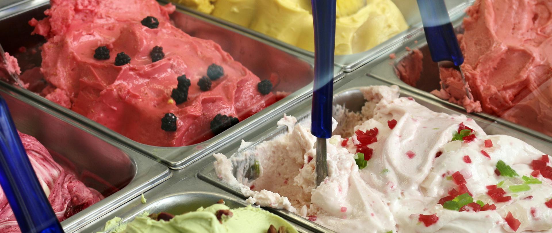 Zdjęcie przedstawia kuwety z lodami różnych smaków. Wbite w nie są również szpatułki do nakładania lodów.