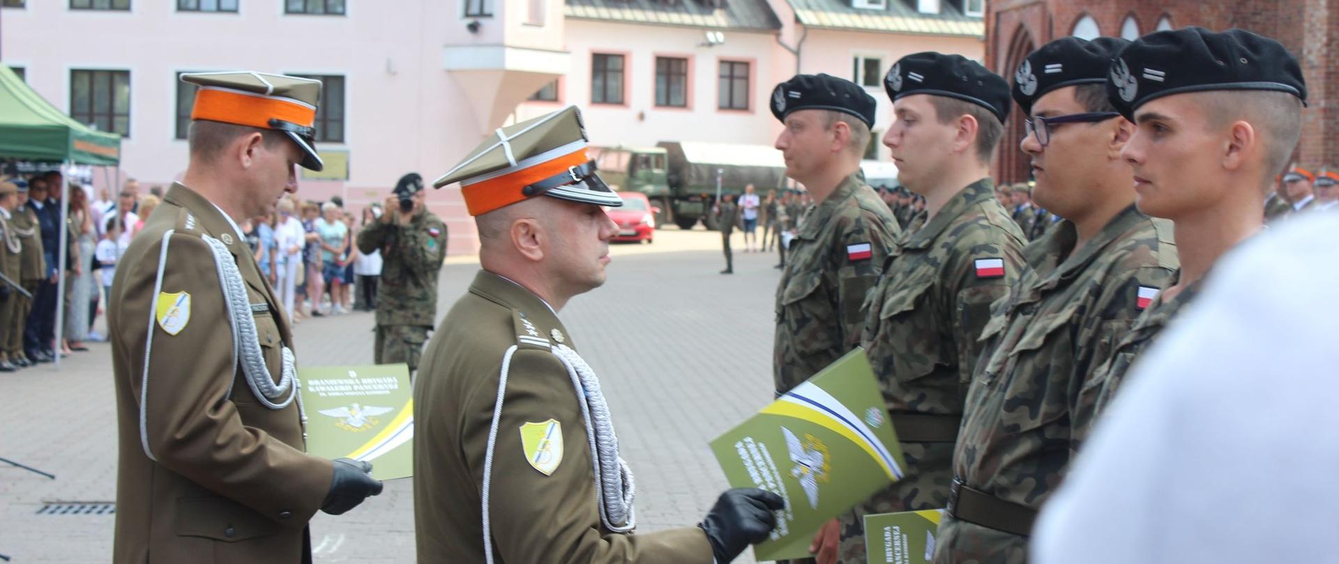 Duży plac w tle budynki. Na placu dowódca wojskowy wręcza awanse. Z prawej w szeregu żołnierze.