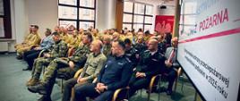 Sala konferencyjna wypełniona uczestnikami w mundurach służb mundurowych i garniturach, siedzącymi w rzędach i słuchającymi wystąpienia; po prawej stronie widoczny ekran z napisem „Państwowa Straż Pożarna”.