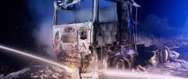 Zdjęcie przedstawia całkowicie spalony ciągnik siodłowy, który stoi na jedni. Pora nocna. Nad samochodem unosi się dym. Na zdjęciu widać również prądy wody. 