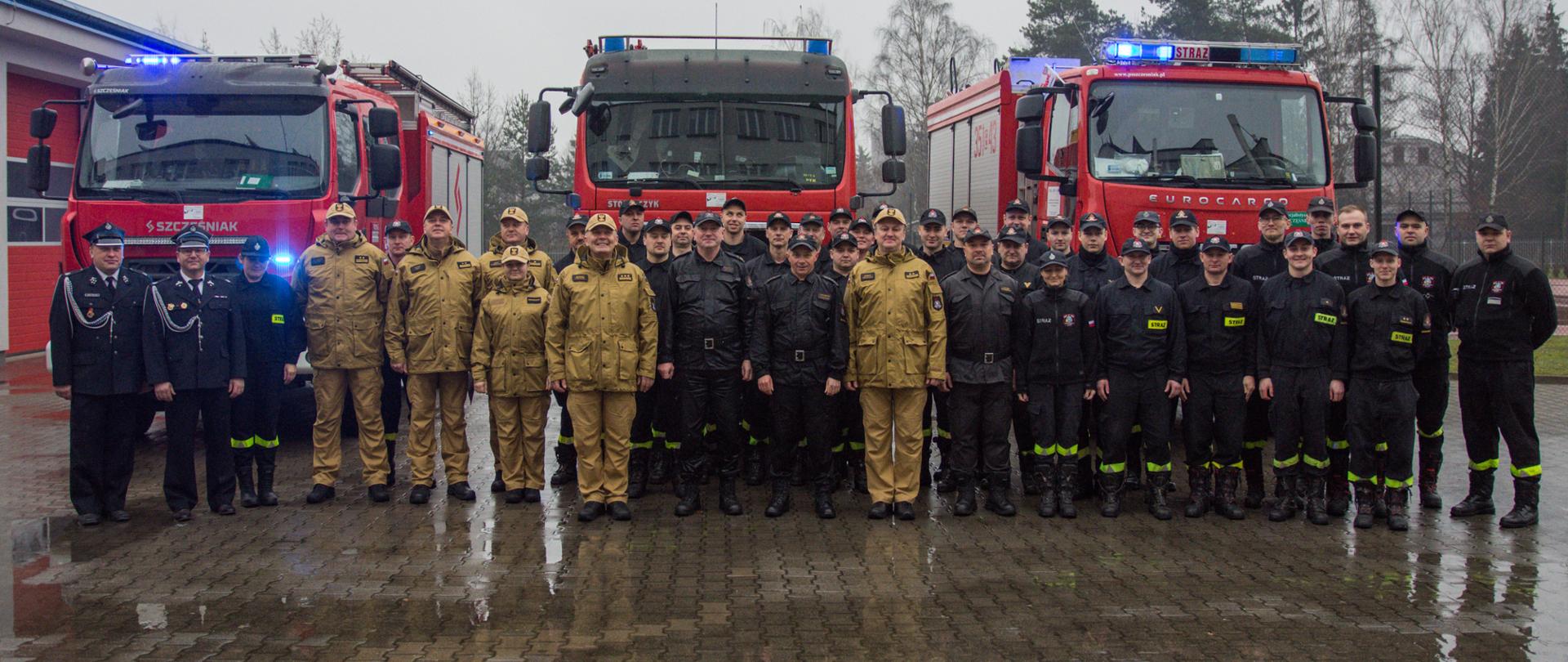 Na zdjęciu strażacy w mundurach koloru czarnego i piaskowego stoją na tle samochodów strażackich.