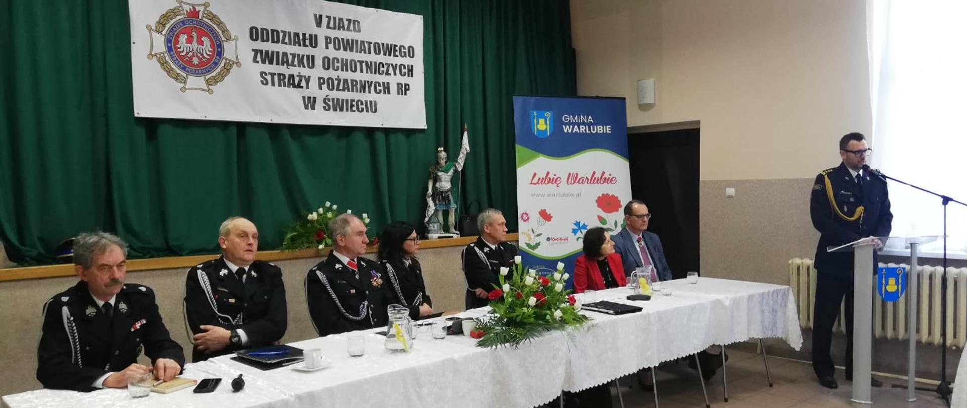 Na zdjęciu widać stół przy którym siedzą druhowie Ochotniczych Straży Pożarnych, Starosta Świecki oraz Wójt Gminy Warlubie wchodzący w skład prezydium Piątego Zjazd Oddziału Powiatowego Związku Ochotniczych Straży Pożarnych RP w Świeciu. W tle po prawej przy mównicy przemawia starszy brygadier Piotr Tkaczyński - Komendant Powiatowy Państwowej Straży Pożarnej w Świeciu.
