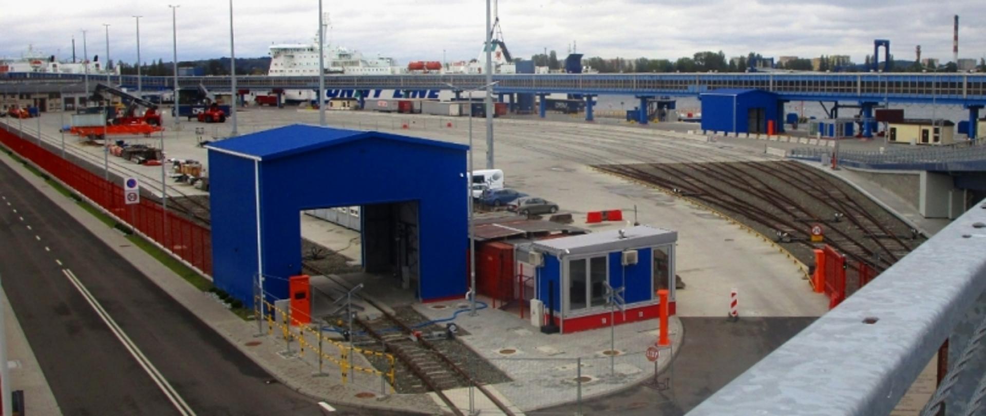 Teren przemysłowy z torami, budynkami i promem w tle, pod pochmurnym niebem.