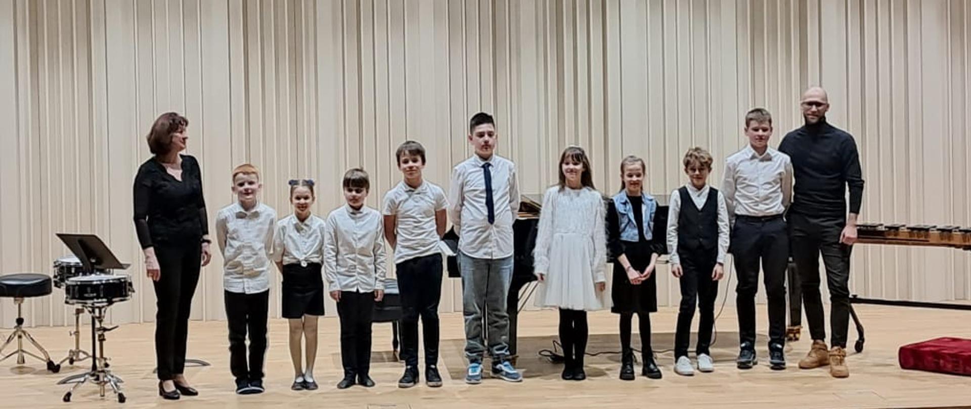 Na zdjęciu grupa odświętnie ubranych uczniów. Po lewej stronie kobieta brunetka ubrana na czarno, a po prawej stronie mężczyzna w okularach ubrany na czarno. Tło obrazka w odcieniach beżu.