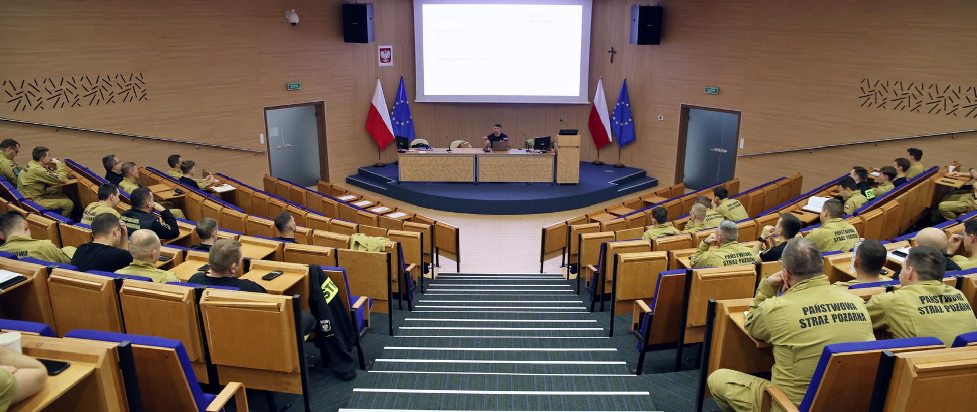 Widok z góry na funkcjonariuszy PSP siedzących w auli podczas wykładu, w głębi na środku przy stole konferencyjnym stojącym na podwyższeniu siedzący prelegent. Za nim rozwinięty ekran multimedialny z wyświetlonym slajdem prezentacji