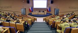 Widok z góry na funkcjonariuszy PSP siedzących w auli podczas wykładu, w głębi na środku przy stole konferencyjnym stojącym na podwyższeniu siedzący prelegent. Za nim rozwinięty ekran multimedialny z wyświetlonym slajdem prezentacji