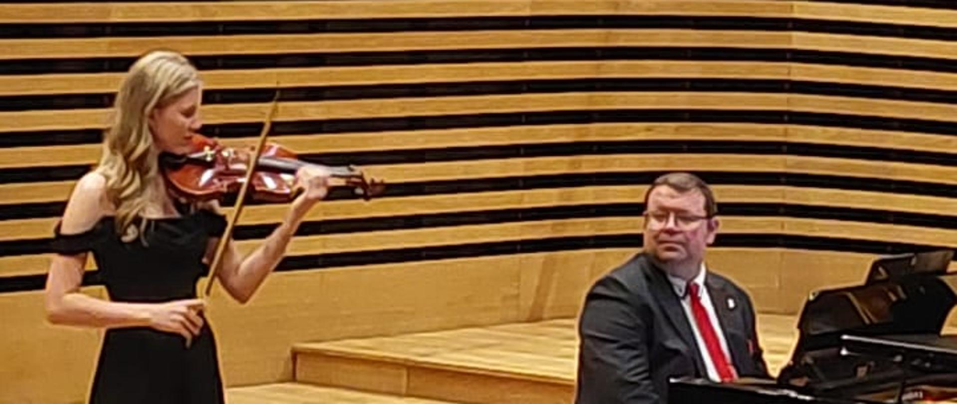 Dziewczyna gra na skrzypcach, mężczyzna akompaniuje na fortepianie.