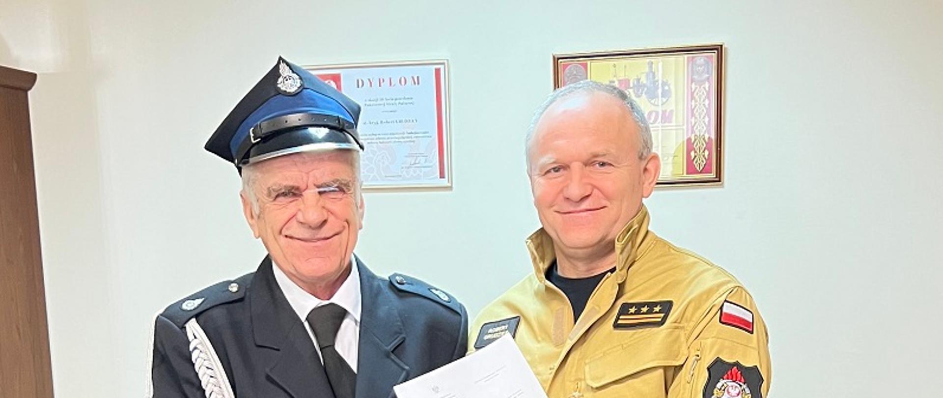 Na zdjęciu widać Komendanta Powiatowego PSP wraz z druhem, któremu wręcza decyzję o przyznaniu świadczenia ratowniczego. Zdjęcie wykonane w gabinecie Komendanta Powiatowego. W tle dwa brązowe fotele. 
