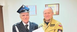 Na zdjęciu widać Komendanta Powiatowego PSP wraz z druhem, któremu wręcza decyzję o przyznaniu świadczenia ratowniczego. Zdjęcie wykonane w gabinecie Komendanta Powiatowego. W tle dwa brązowe fotele. 