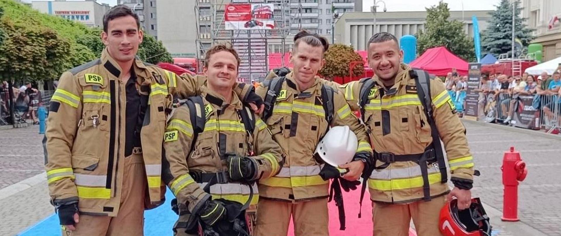 Zdjęcie wykonane podczas zawodów Najtwardszy Strażak. Na fotografii grupowe zdjęcie czterech strażaków z komendy powiatowej Państwowej Straży Pożarnej w Mielcu podczas zawodów w Rzeszowie. Są oni ubrani w umundurowania specjalne bojowe z hełmami strażackimi w ręku. Zdjęcie wykonane podczas zawodów przed budynkiem Urzędu Wojewódzkiego w Rzeszowie." 