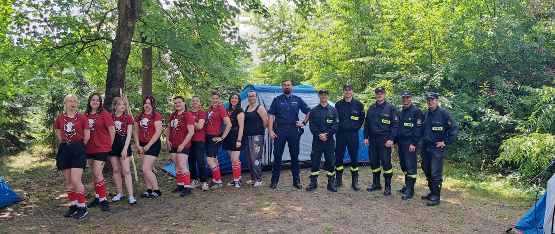 harcerki i funkcjonariusze straży pożarnej i policji stoją na polanie w lesie, w tle drzewa i rozstawione namioty