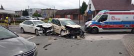 Widoczne rozbite przody dwóch pojazdów uczestniczących w zdarzeniu. Obok karetka pogotowia oraz samochód Policyjny,. Ja jezdni widoczna duża ilość elementów karoserii samochodowej oraz wycieki płynów eksploatacyjnych zabezpieczone częsciowo sorbentem. Obok aut stoją strażacy prowadząc czynności
