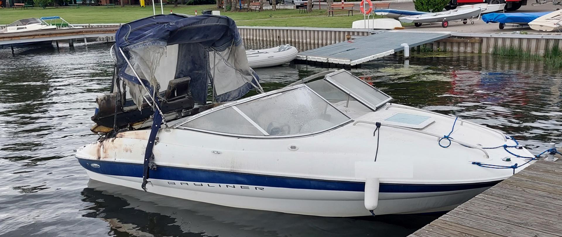 Zdjęcie przedstawia łódź motorową przycumowaną do pomostu. Tył łodzi w okolicach silnika uległ spaleniu. Na zdjęciu widać również plażę, samochód strażacki oraz inne łodzie.