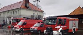Na zdjęciu widać samochody straży pożarnej przy budynku Gminy w Żelazkowie. Pogoda deszczowa, zachmurzenie duże. Wśród 4 samochodów w kolorze czerwonym samochód dowodzenia i łączności z rozłożonymi antenami.
