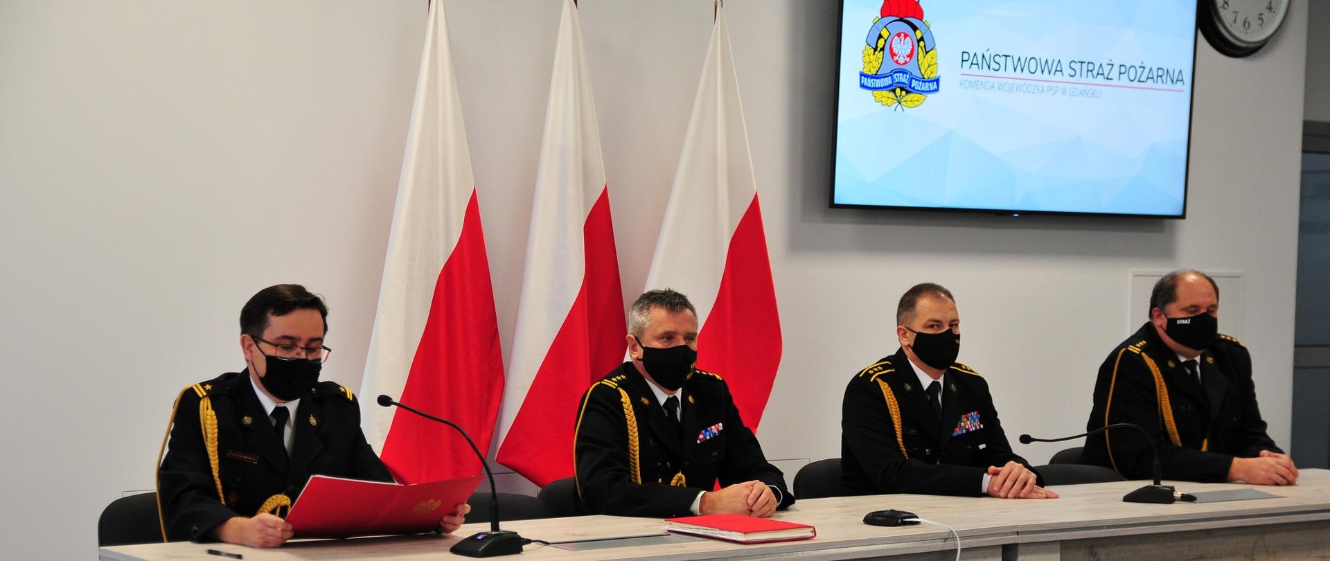 Wideokonferencja z Wojewodą Pomorskim i Pomorskim Komendantem Wojewódzkim PSP - Komendant Wojewódzki wraz z Zastępcami przy stole podczas wideokonferencji. W tle flagi państwowe.