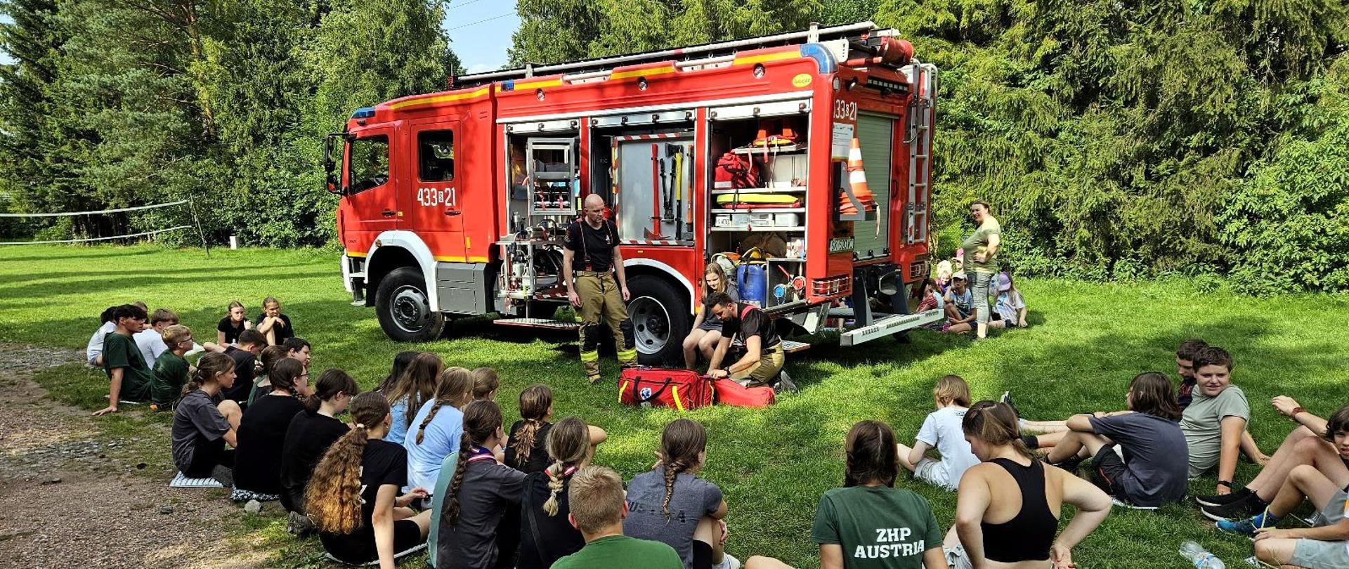 W lesie przy samochodzie specjalnym ratownictwa technicznego, wokół niego siedzi grupa dzieci, przy samochodzie strażacy, którzy prezentują wyposażenie samochodu.