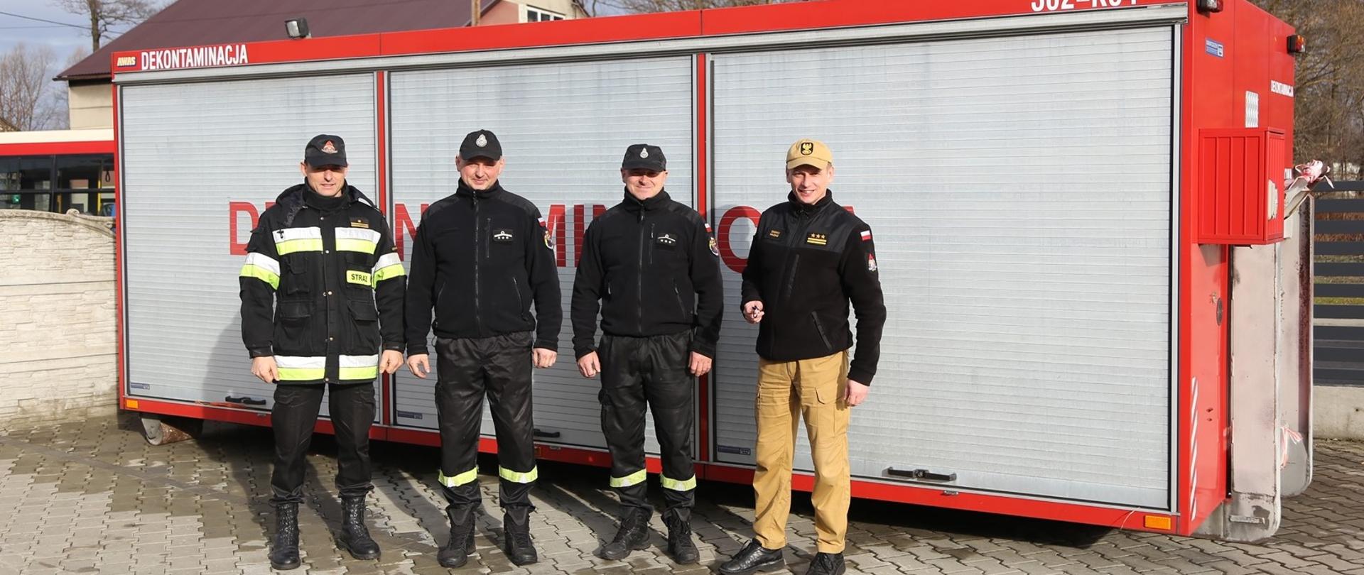 Zastępca Komendanta Centralnej Szkoły PSP w towarzystwie dwóch druhów OSP Zawiercie-Marciszów oraz oficera CS PSP na tle przekazywanego kontenera dekontaminacyjnego