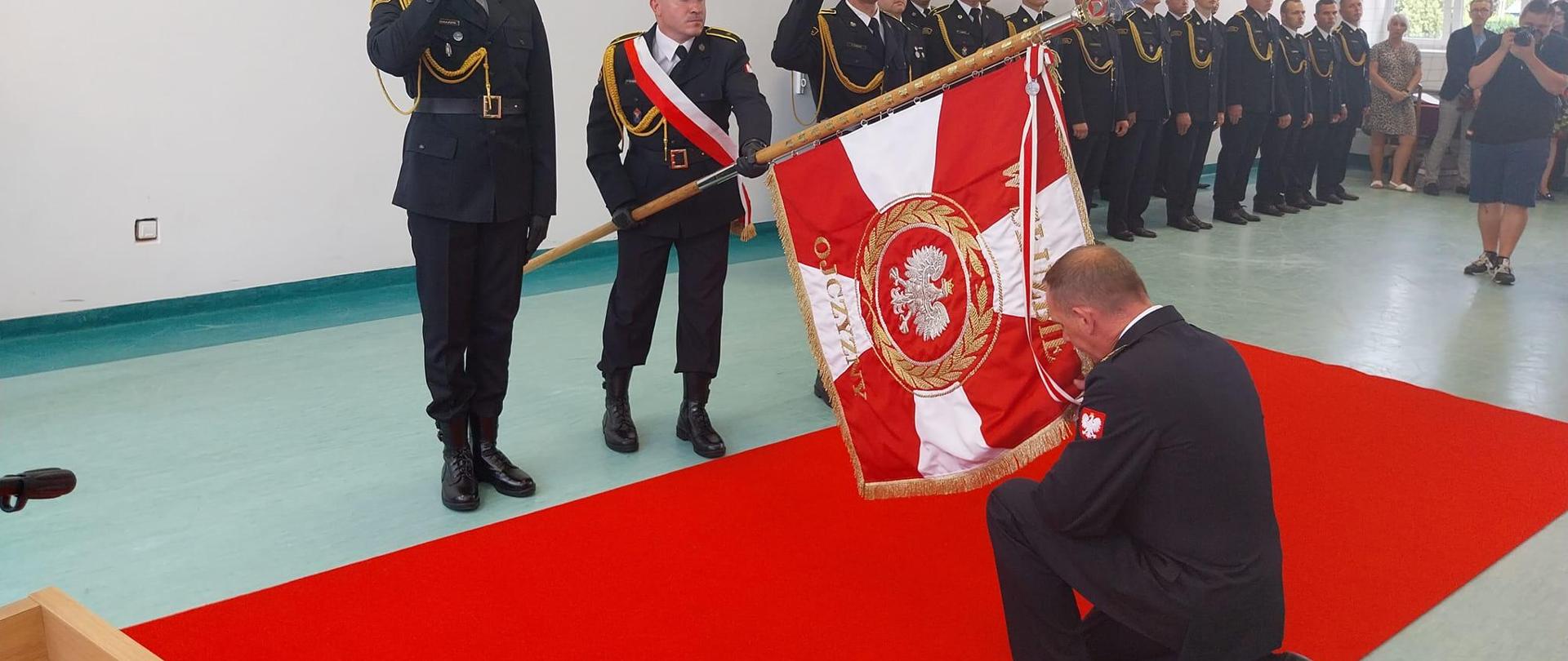 Na zdjęciu widać grupę osób strażaków w mundurach oraz osoby cywilne. Trzech strażaków w czapkach stoi przed czerwonym dywanem. Strażak w środku ma szarfę biało-czerwoną oraz trzyma sztandar. Naprzeciwko ich na czerwonym dywanie na jedno kolano klęka strażak bez czapki na głowie. Całuje sztandar, który jest przechylony w jego stronę. W tle widać pozostałe osoby. Uroczystość odbywa się na sali gimnastycznej. Jest widno.