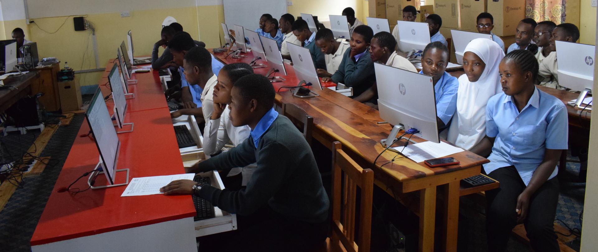 Młodzież siedząca w ławkach przed ekranami komputerów.