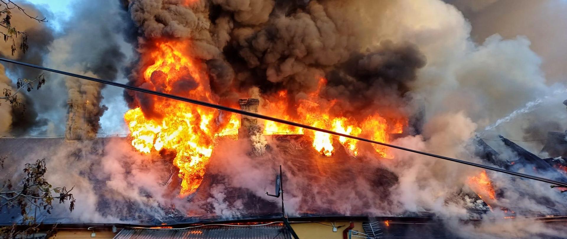 Widoczny dach palącego się budynku, duże płomienie oraz kłęby czarnego dymu