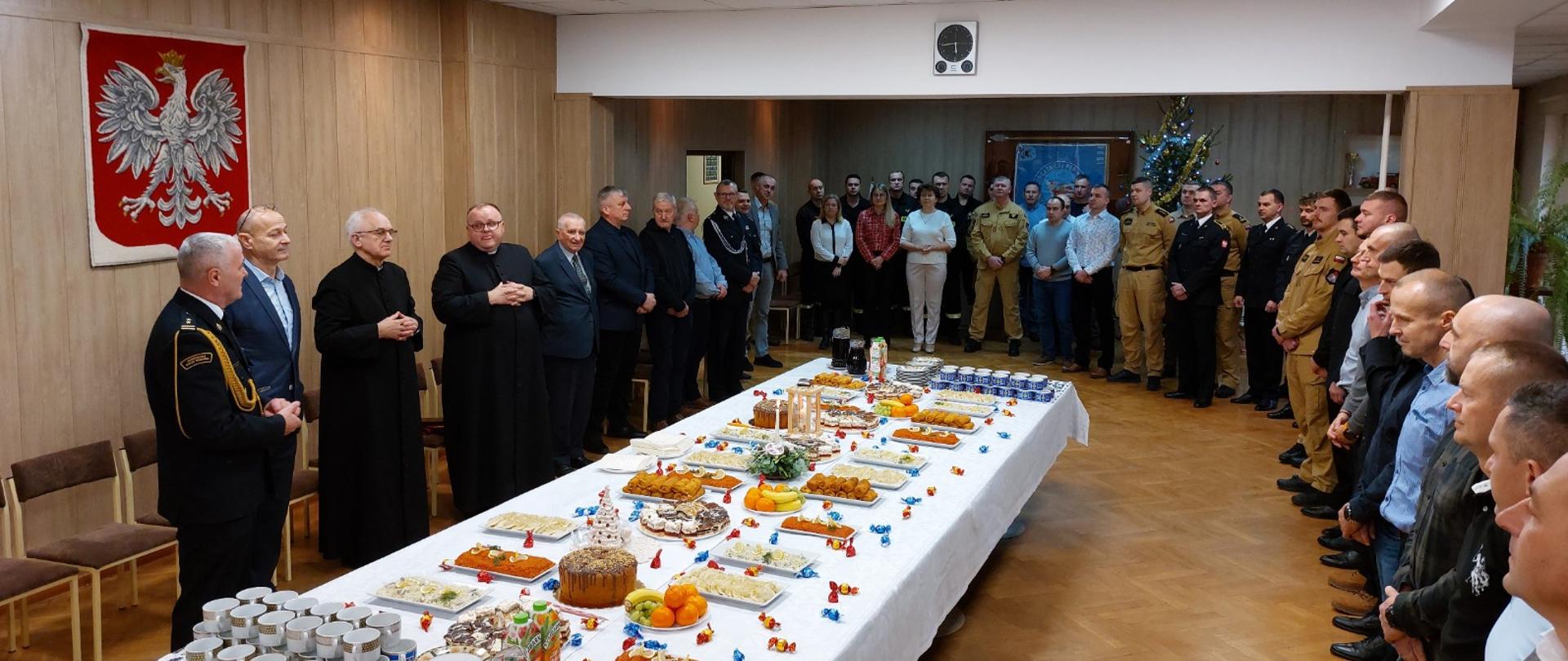 Uroczyste wigilijne spotkanie opłatkowe w sali reprezentacyjnej – przy długim stole nakrytym białym obrusem, zastawionym ciastami, owocami i napojami, stoją zgromadzeni uczestnicy. Po obu stronach sali ustawieni są strażacy PSP w mundurach wyjściowych, pracownicy cywilni oraz zaproszeni goście, w tym duchowni. Na ścianie widoczny jest godło Polski, a w tle ubrana choinka.